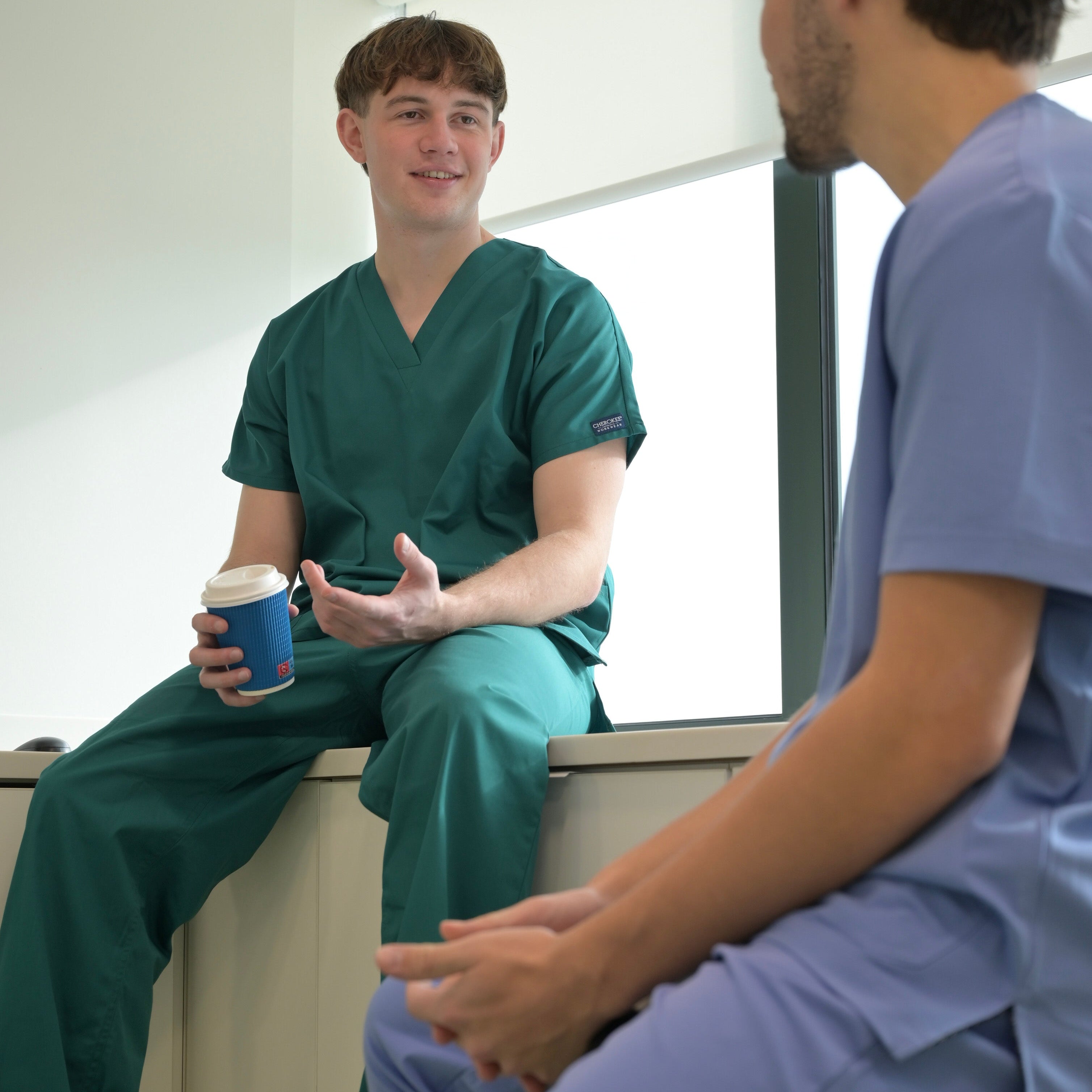 Deux dentistes qui discutent dans un cabinet, portant des blouses médicales Koi de couleur bleu ciel et vert forêt