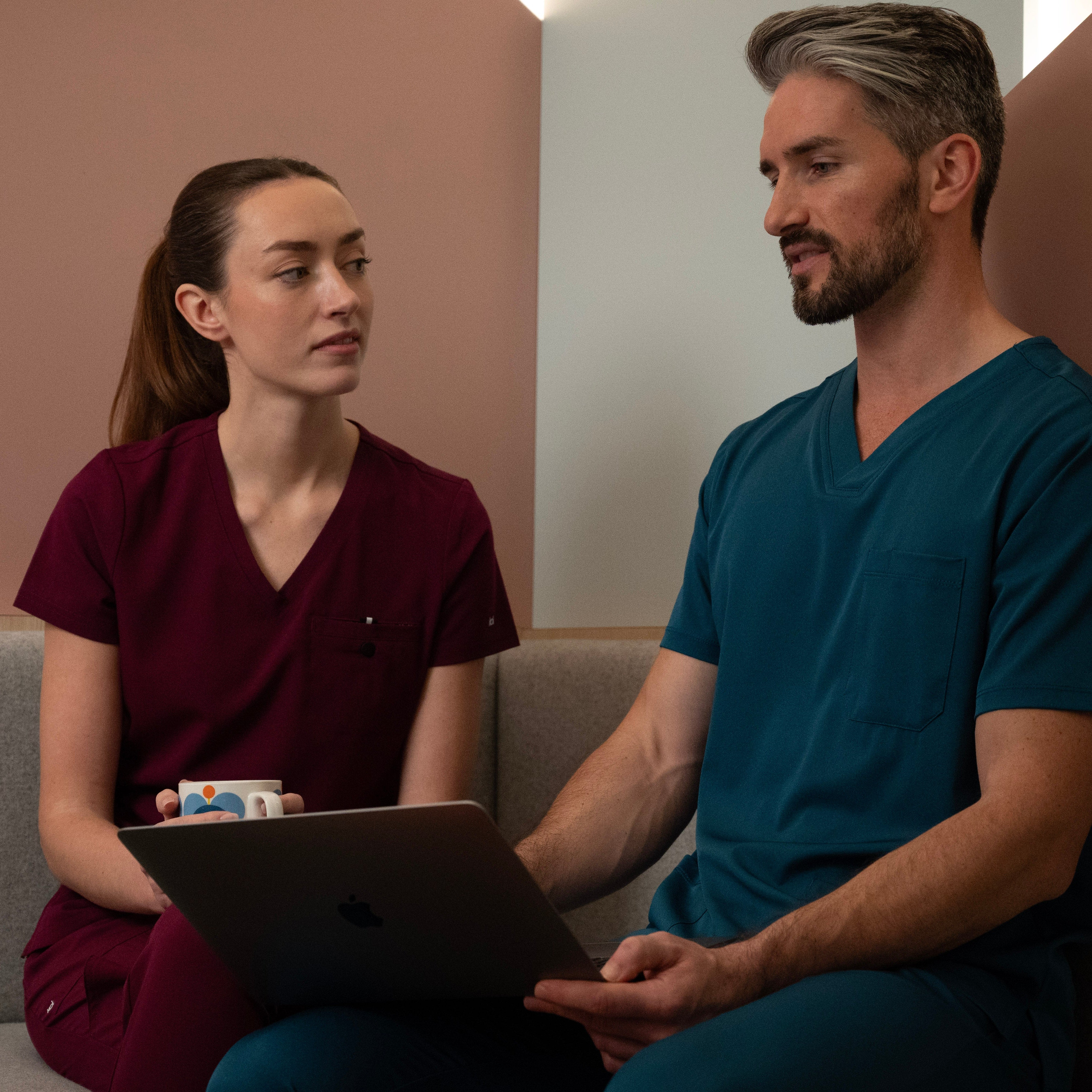 Deux soignants en uniforme, l’un portant une blouse Koi Next Gen bordeaux Prête à travailler et l’autre une blouse Healing Hands bleu caraïbes.