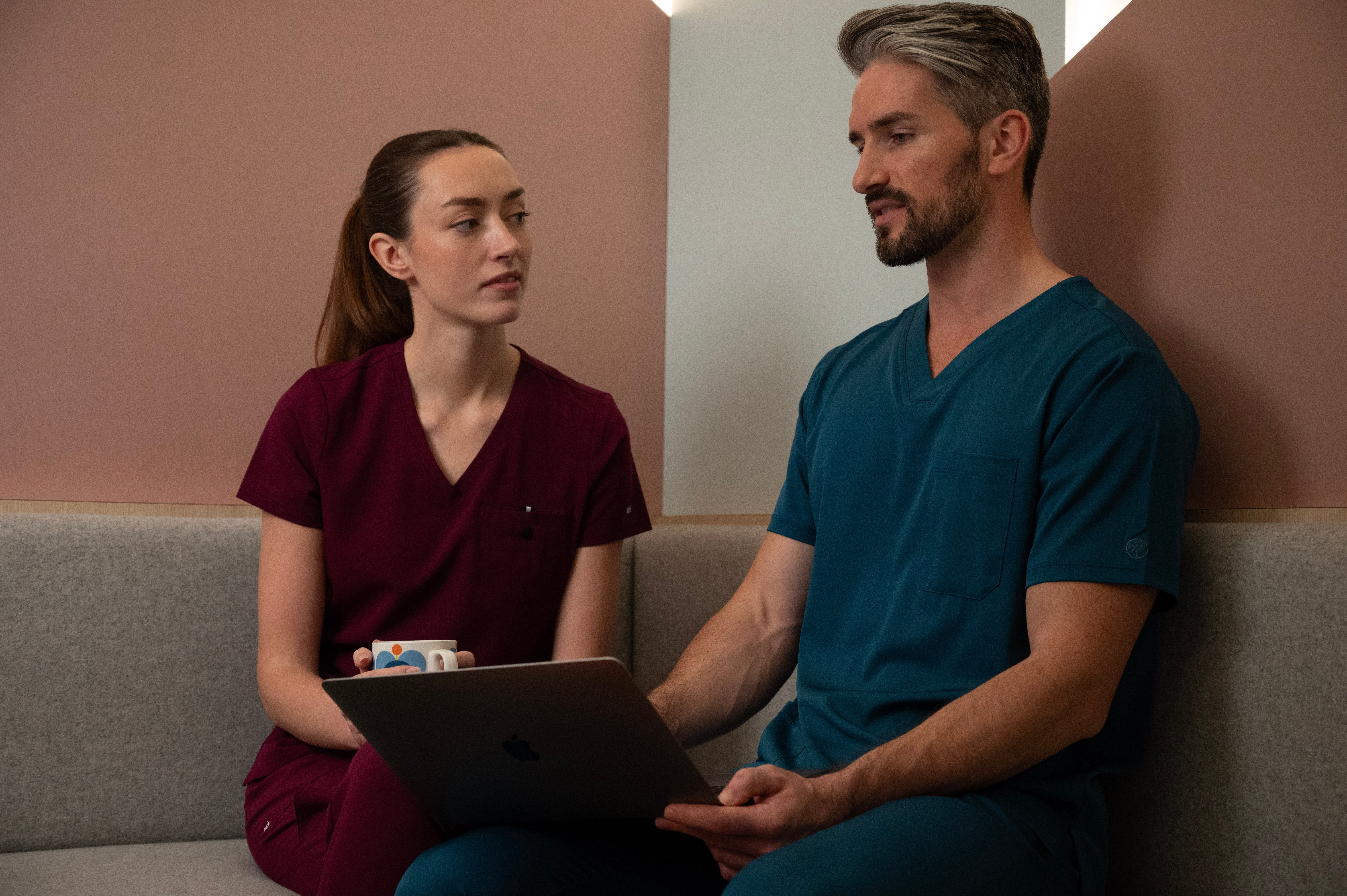 Deux soignants en uniforme, l’un portant une blouse Koi Next Gen bordeaux Prête à travailler et l’autre une blouse Healing Hands bleu caraïbes. 